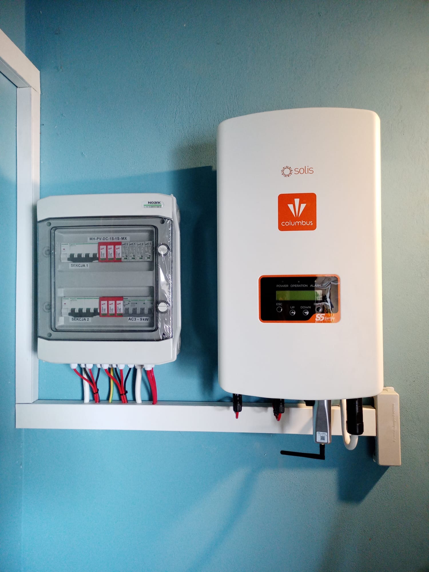 Biała skrzynka rozdzielcza z zabezpieczeniami i wyprowadzonymi kablami oraz biały inwerter Solis Columbus S5 Series zamontowane na ścianie o błękitnym kolorze.