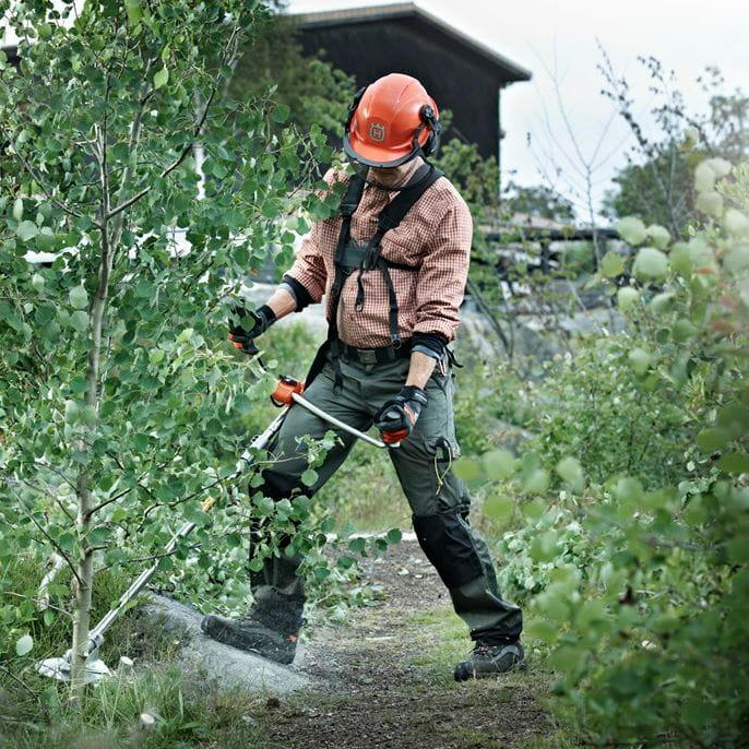 Pracownik w kasku Husqvarna i odzieży ochronnej, w trakcie wycinania krzewów przy użyciu specjalistycznego narzędzia w terenie zielonym w Warszawie.
