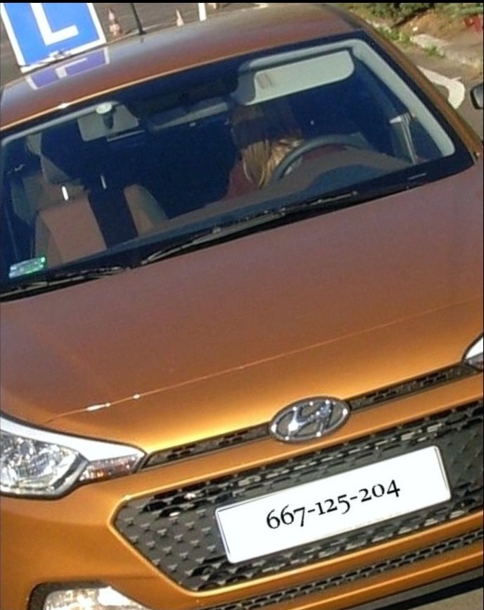 Przód pomarańczowego samochodu z tablicą z numerem telefonu i znakiem 'L' na dachu, widoczna osoba za kierownicą.