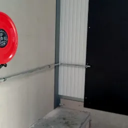 Czerwona zwijana gaśnica hydrantowa na ścianie z szarą rurą doprowadzającą wodę, widoczne złącza i zawór, tło stanowi ściana z blachy trapezowej i czarna płyta.