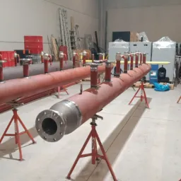 Długie, stalowe kolektory rurowe z licznymi króćcami, pomalowane na czerwono i szaro, ustawione na czerwonych stojakach w hali produkcyjnej.