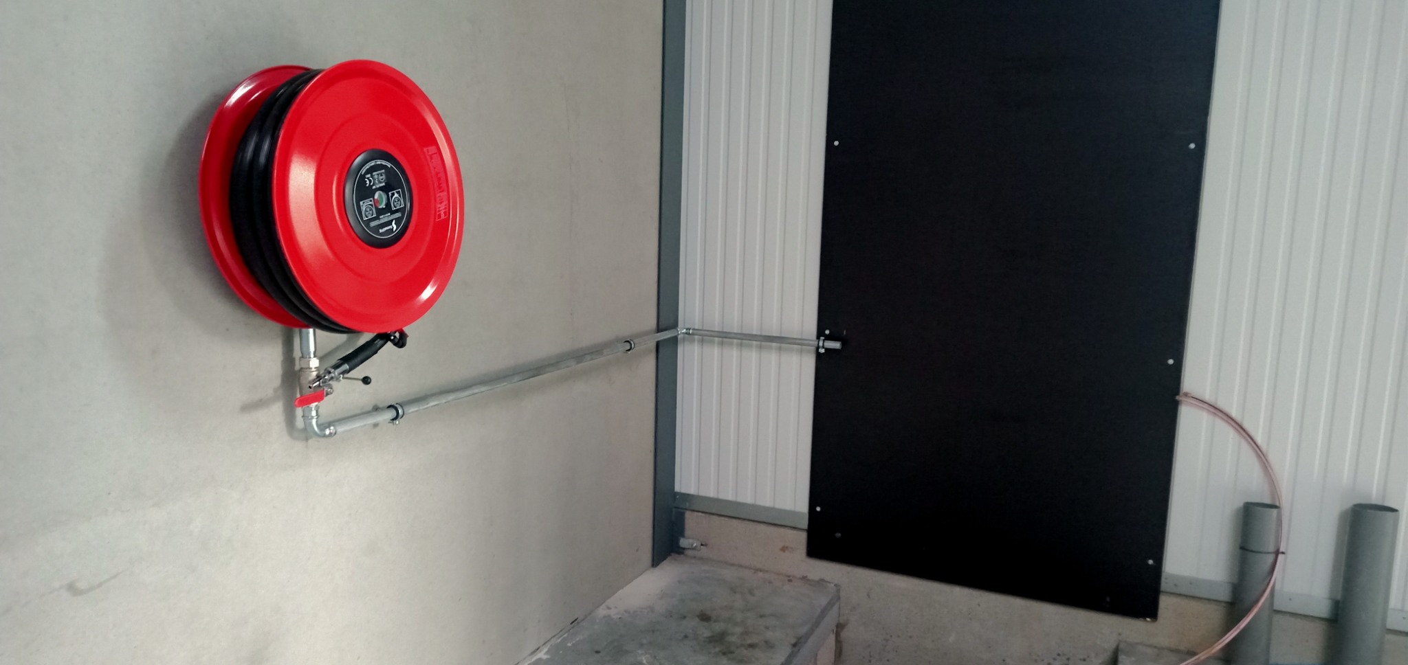 Czerwona zwijana gaśnica hydrantowa na ścianie z szarą rurą doprowadzającą wodę, widoczne złącza i zawór, tło stanowi ściana z blachy trapezowej i czarna płyta.