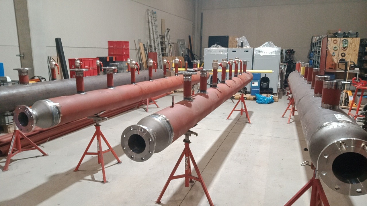Długie, stalowe kolektory rurowe z licznymi króćcami, pomalowane na czerwono i szaro, ustawione na czerwonych stojakach w hali produkcyjnej.