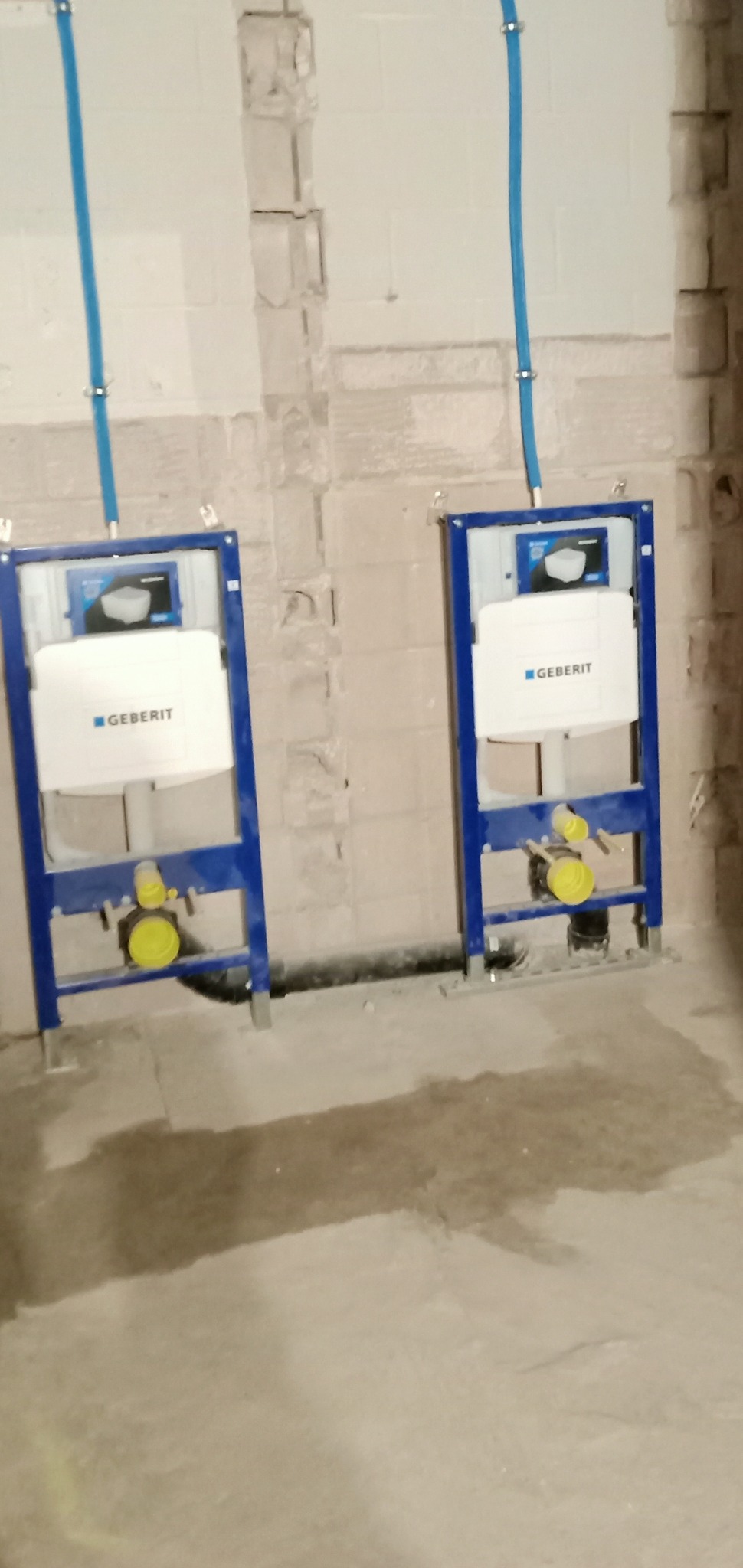 Dwa podtynkowe stelaże WC marki Geberit w nieukończonej łazience, z widocznymi nieotynkowanymi ścianami i wystającymi rurami doprowadzającymi wodę.