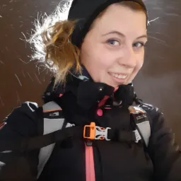 Uśmiechnięta kobieta w czapce i kurtce sportowej z plecakiem, patrząca w obiektyw, oświetlona od tyłu, na ciemnym tle.