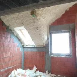Poddasze w trakcie ocieplania pianką PUR, widoczne ceglane ściany, okno połaciowe i okno pionowe, fragmenty pianki na podłodze.