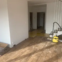 Pokój w trakcie prac wykończeniowych: częściowo ułożone panele podłogowe, ślady stóp, odkurzacz i kartony paneli w rogu pomieszczenia.