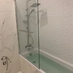 Nowoczesna łazienka z wanną, szklanym parawanem i armaturą w kolorze chromu, wykończona białymi płytkami o zróżnicowanej fakturze na ścianach. Woda w wannie lekko zabrudzona.