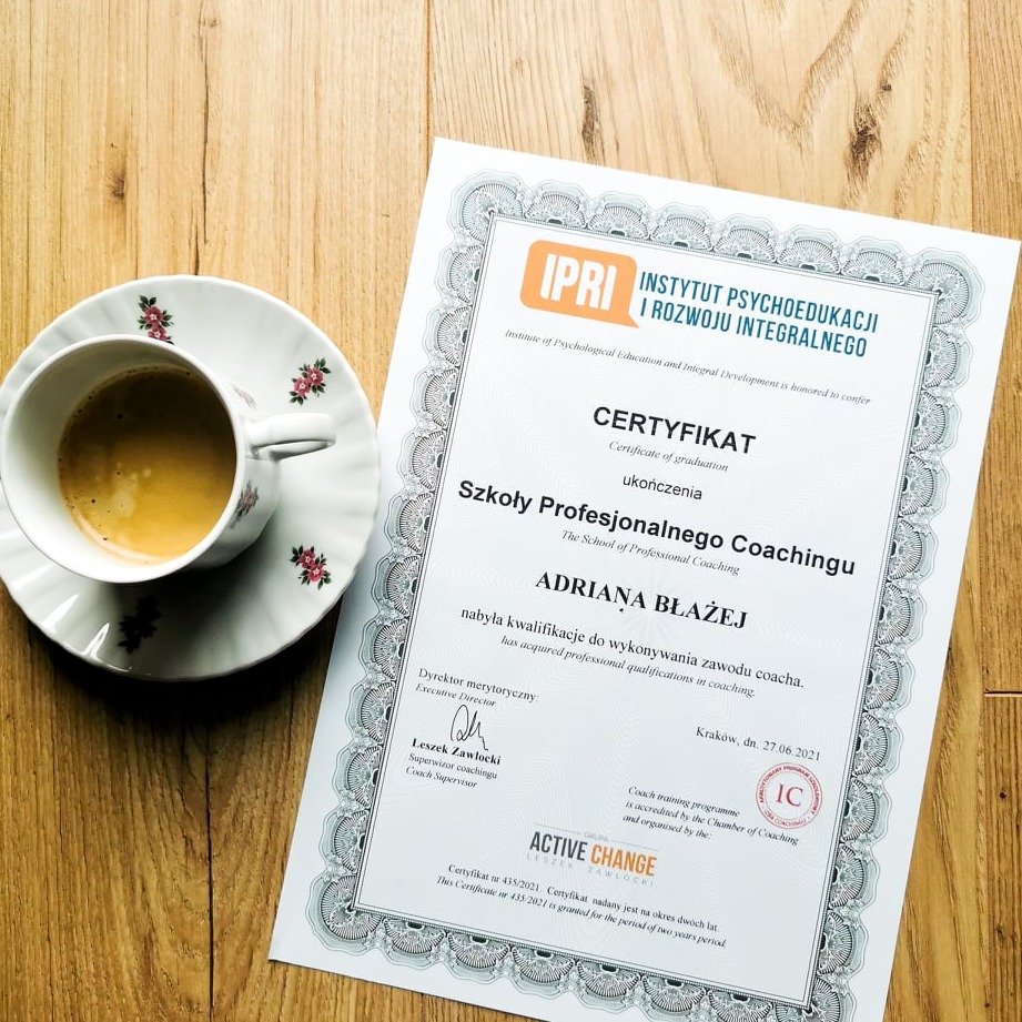 Certyfikat ukończenia Szkoły Profesjonalnego Coachingu Adriana Błażej, leżący obok filiżanki kawy na drewnianym stole.