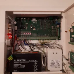 Centrala alarmowa z akumulatorem i transformatorem w białej obudowie, zainstalowana w Pile, widoczne liczne przewody i elementy elektroniczne.