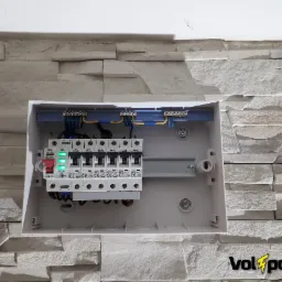 Rozdzielnica elektryczna z bezpiecznikami, zamontowana na ścianie z dekoracyjnej cegły, z widocznymi przewodami i listwą zaciskową, z włączonymi zielonymi diodami.