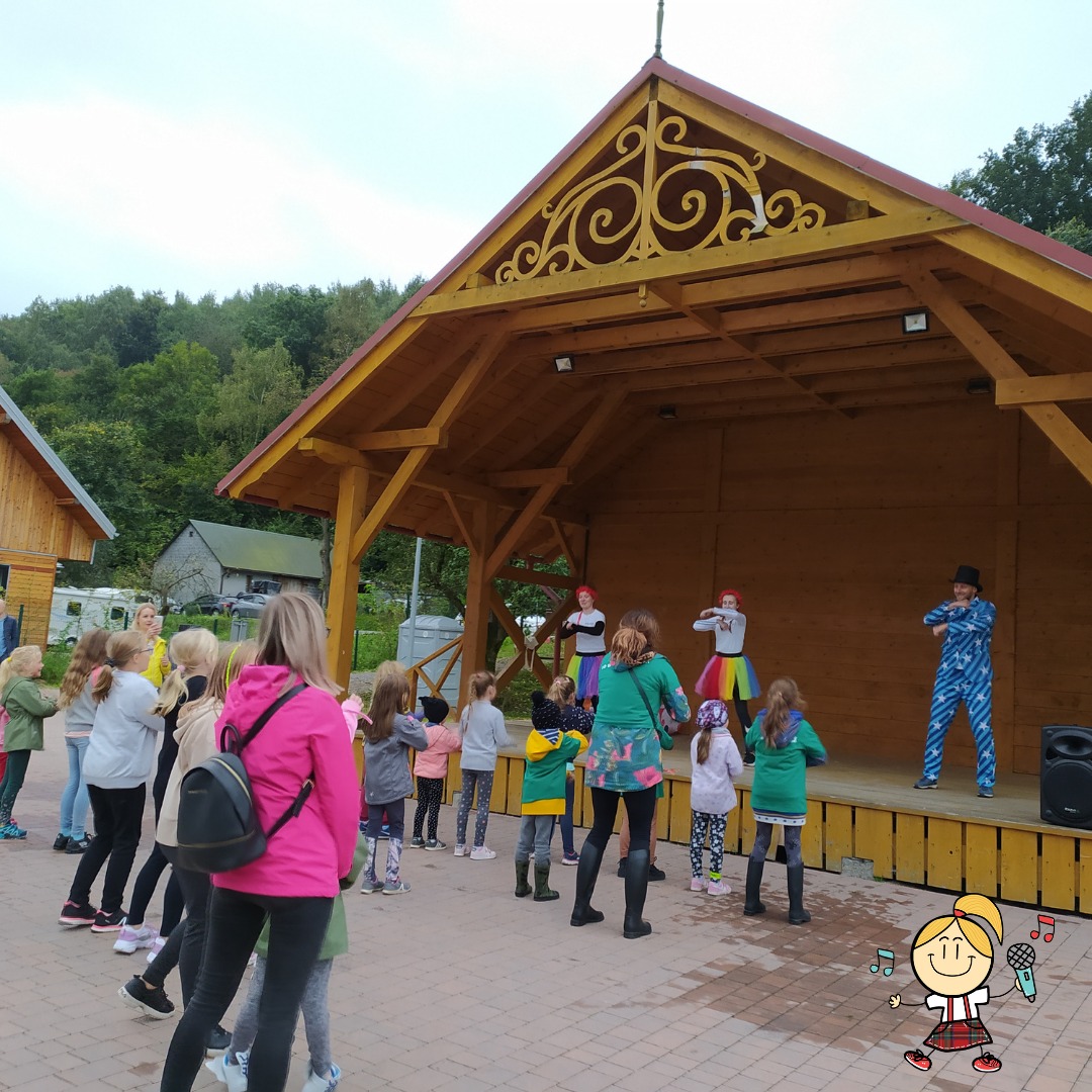Animator w niebieskim kostiumie w gwiazdki i czarnym cylindrze prowadzi taniec dla grupy dzieci na drewnianej scenie z ozdobnym szczytem, na zewnątrz.