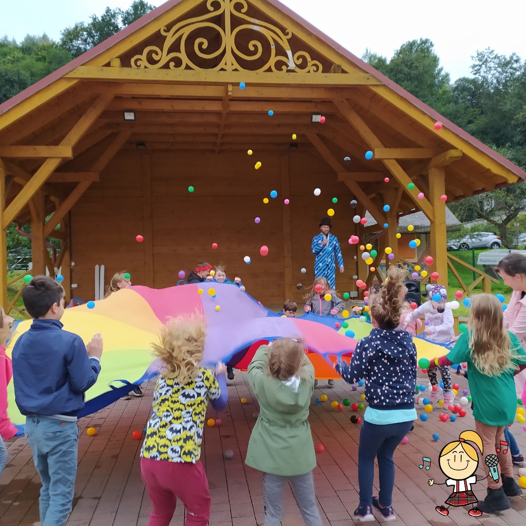 Dzieci bawiące się pod kolorową chustą animacyjną, z góry spadają kolorowe piłeczki, w tle drewniana scena z animatorem w niebieskim kostiumie.