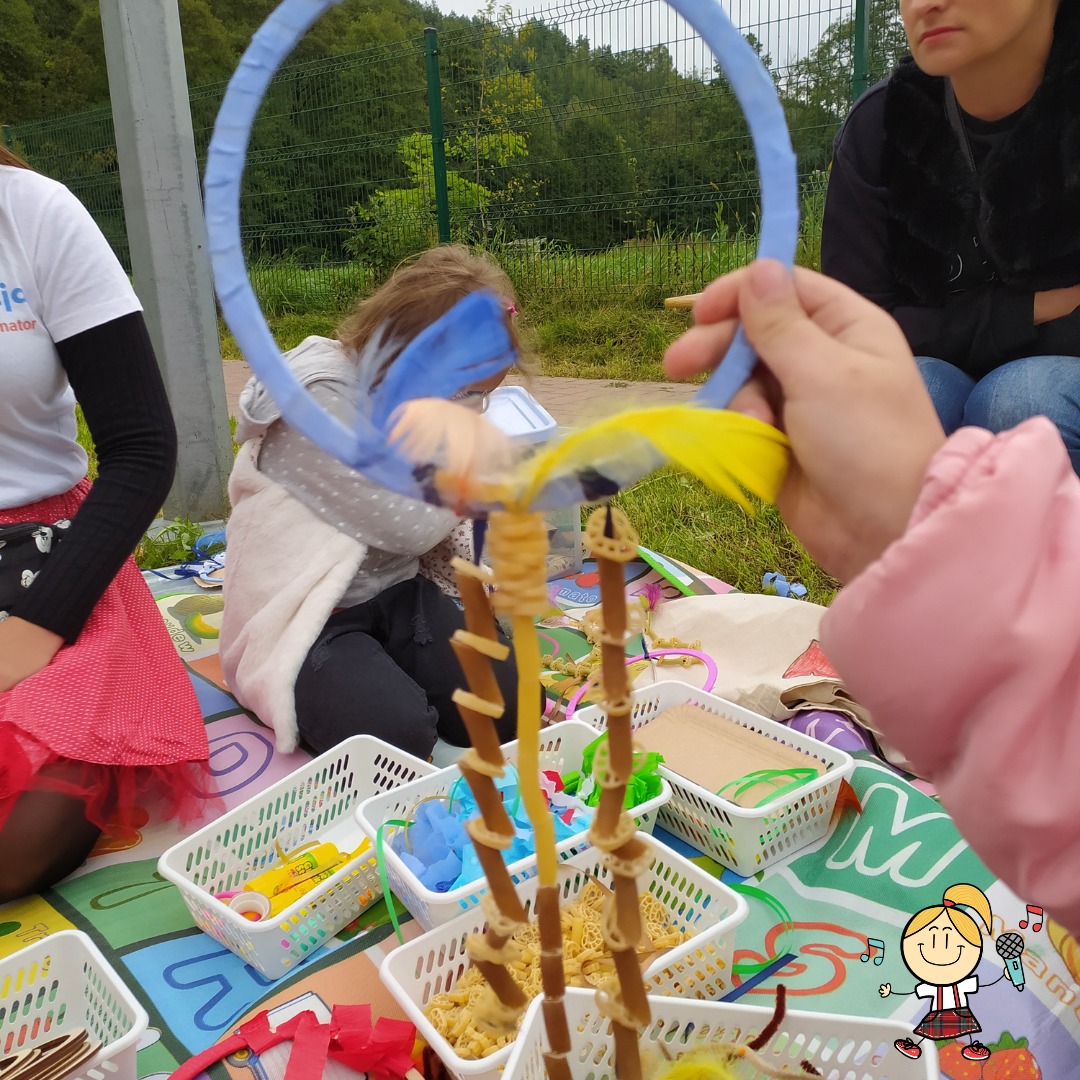 Dziecko trzyma niebieskie kółko z piórami, na pierwszym planie koszyczki z makaronem i materiałami plastycznymi, w tle siedzące osoby na kocu.
