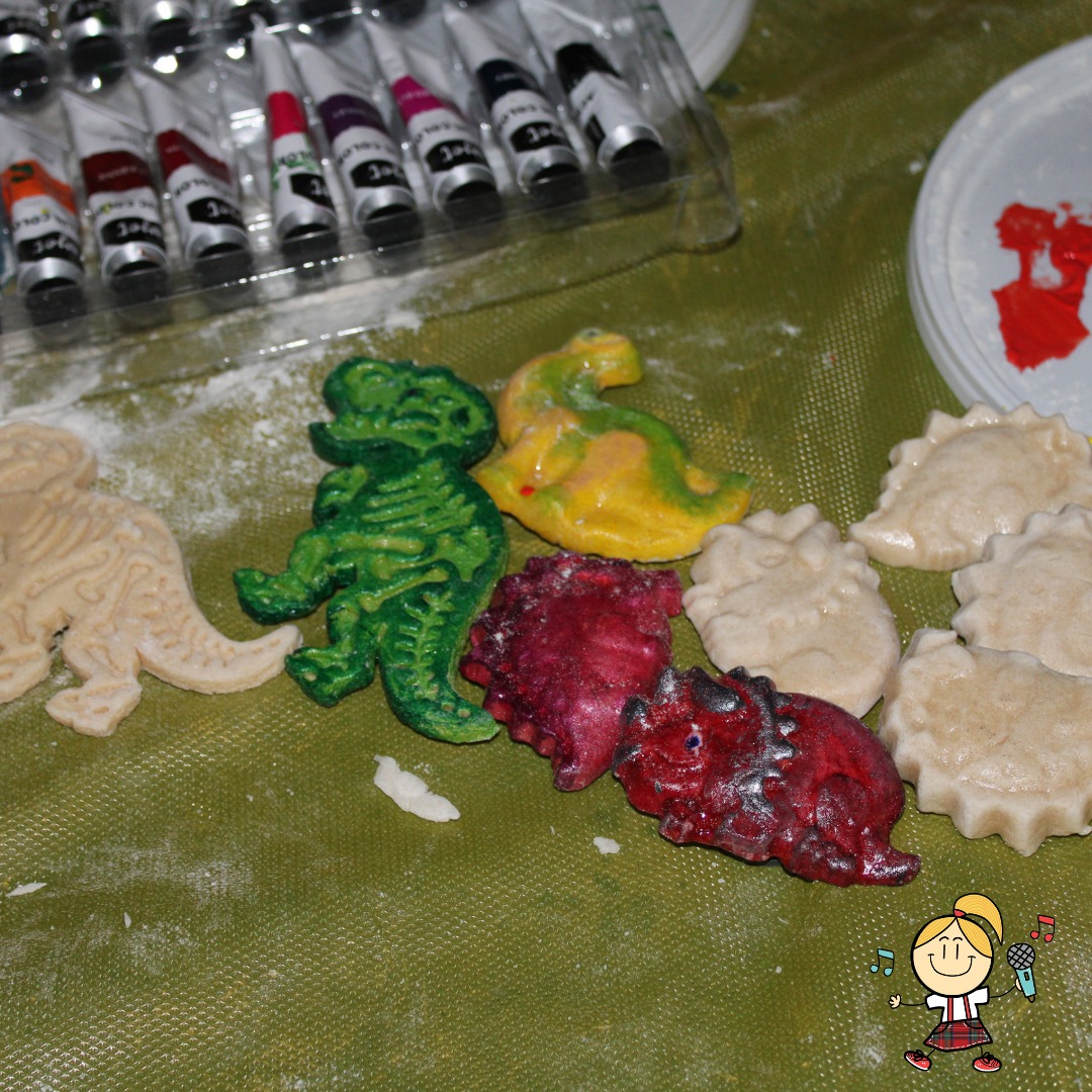 Kolorowe figurki dinozaurów z masy solnej leżące na zielonym tle, obok tubki z farbami i talerzyk z resztkami farby.