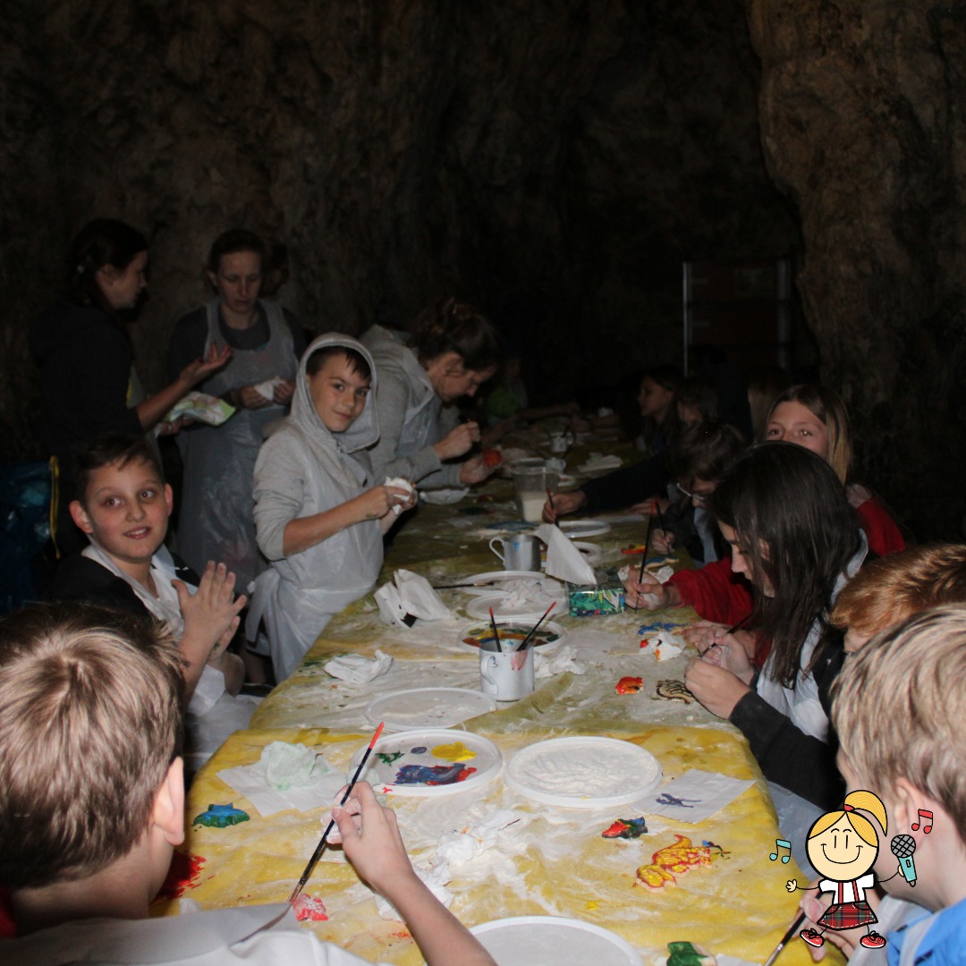 Dzieci malują gipsowe figurki na żółtym stole w jaskini, ubrani w foliowe fartuchy, widoczny pędzel, farby i gotowe pomalowane figurki.