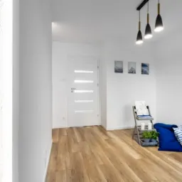 Jasne wnętrze z drewnianą podłogą, białą ścianą z dekoracyjną strukturą i oświetleniem LED, drzwi z przeszkleniami i minimalistycznym wystrojem.