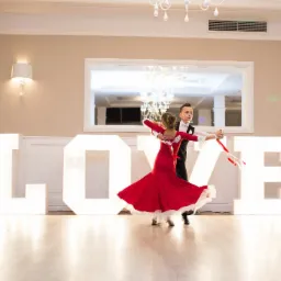 Para młodych tancerzy w strojach galowych tańczy taniec towarzyski na tle podświetlanych liter LOVE podczas eventu.