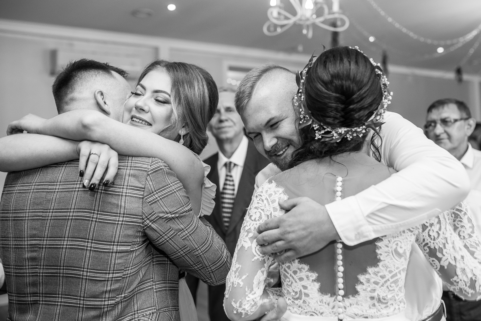 Czarno-białe zdjęcie uśmiechniętej panny młodej w koronkowej sukni, obejmowanej przez gości weselnych w sali z żyrandolem.