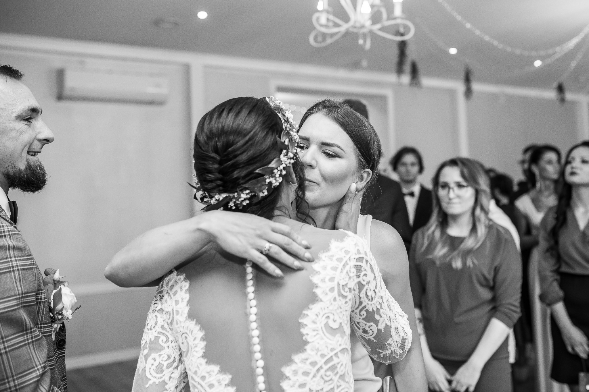Czarno-białe zdjęcie panny młodej w sukni z koronkowymi rękawami, obejmowanej przez kobietę z czułym wyrazem twarzy, w tle goście weselni i kryształowy żyrandol.