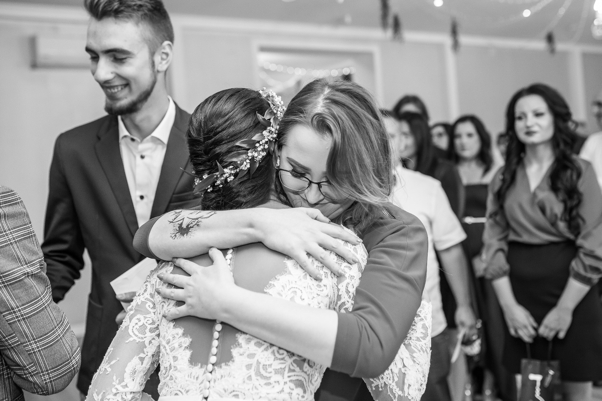 Czarno-białe zdjęcie: Panna młoda w koronkowej sukni z ozdobą we włosach, obejmująca wzruszoną kobietę w okularach na tle gości weselnych.