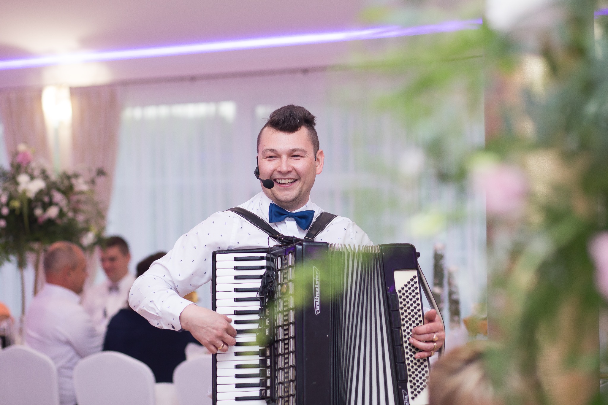 Uśmiechnięty mężczyzna w muszce i koszuli w drobne wzorki gra na akordeonie z mikrofonem nagłownym, w tle siedzący goście weselni i dekoracje kwiatowe.