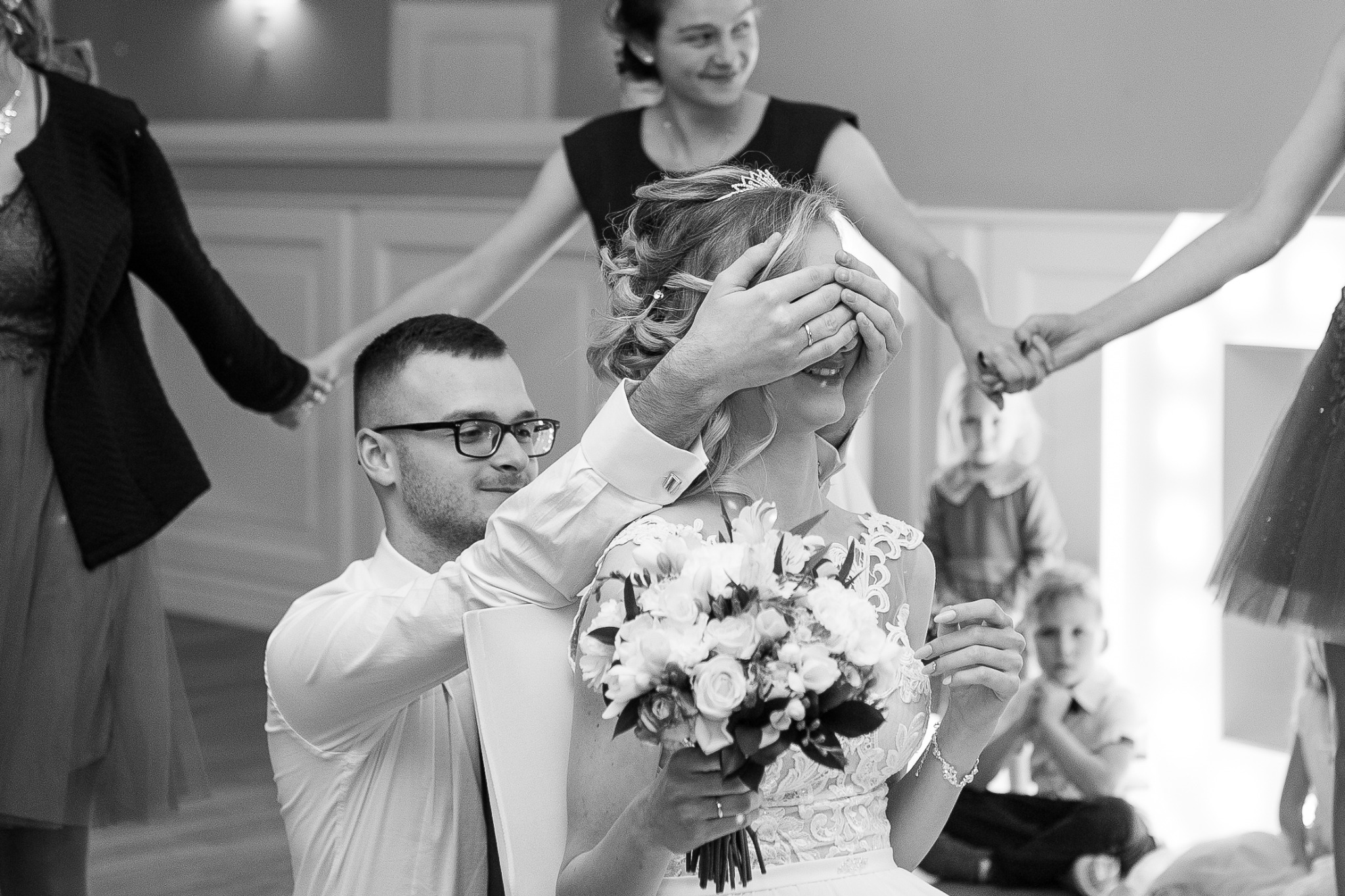 Czarno-białe zdjęcie panny młodej z bukietem, której oczy zasłania mężczyzna w okularach, w tle dzieci i kobiety trzymające się za ręce.