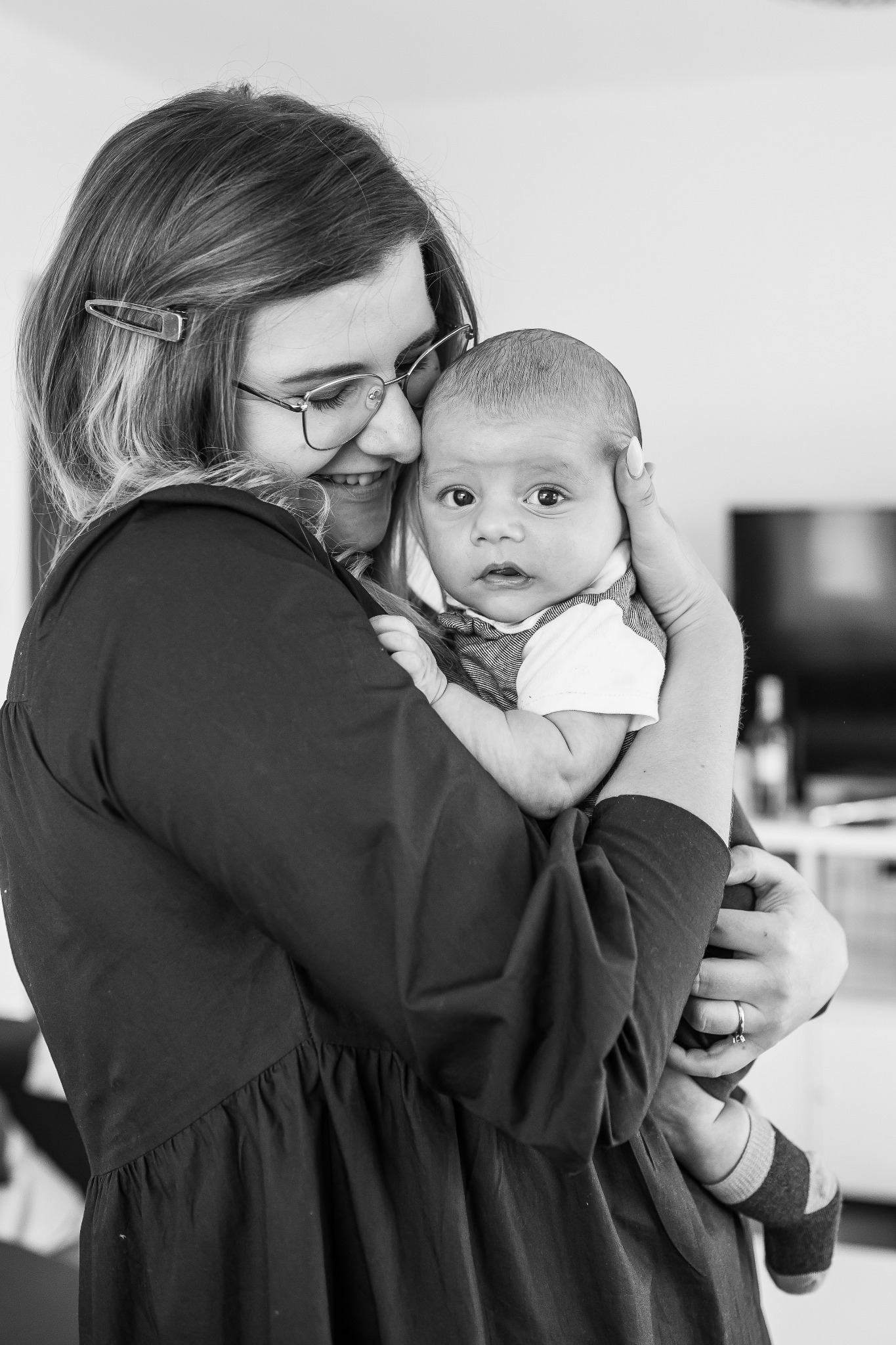 Czarno-białe zdjęcie uśmiechniętej kobiety w okularach trzymającej na rękach niemowlę ubrane w koszulę i sweterek.