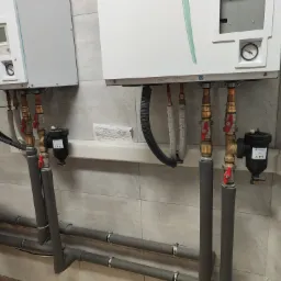 Dwa białe piece wiszące na ścianie wyłożonej szarymi płytkami, z widocznymi rurami, zaworami, filtrami i izolacją termiczną, tworzące system grzewczy w pomieszczeniu.