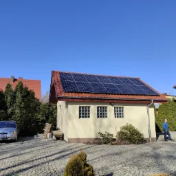 Panele słoneczne na dachu budynku gospodarczego z ceglastą dachówką, na posesji z kostki brukowej, w tle samochód i zieleń.