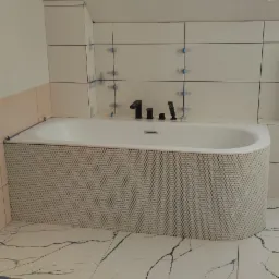 Biała wanna z obudową z mozaiki heksagonalnej, czarna armatura, białe płytki ścienne z niebieskimi krzyżykami dystansowymi, płytki podłogowe imitujące marmur.