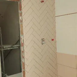 Montaż beżowych płytek ułożonych w jodełkę na ścianie łazienki, widoczne przygotowane przyłącza wodne, fragment podłogi z białych płytek imitujących marmur z czarnymi żyłami, butelka wody...