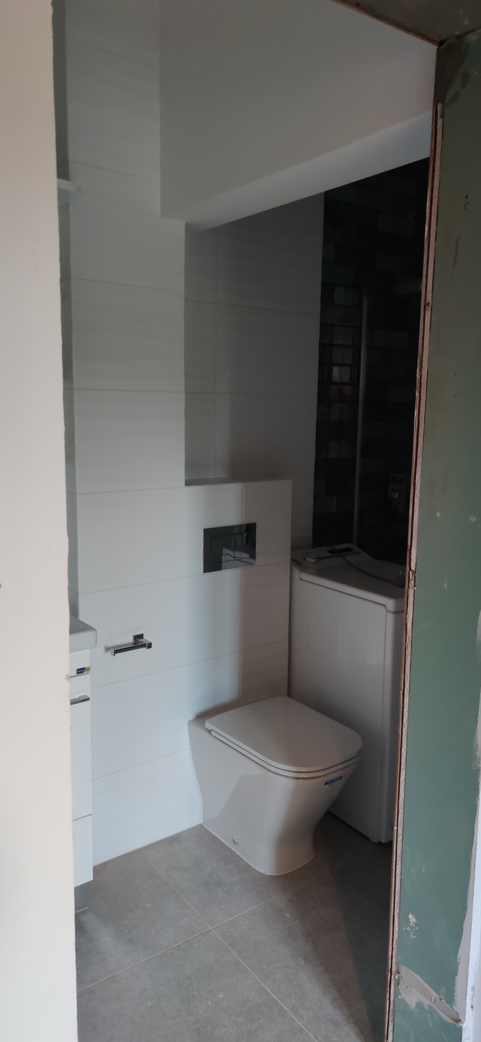 Wnętrze łazienki w trakcie wykańczania: białe płytki na ścianach, wisząca toaleta, szara podłoga, pralka w tle, surowa ościeżnica drzwi.