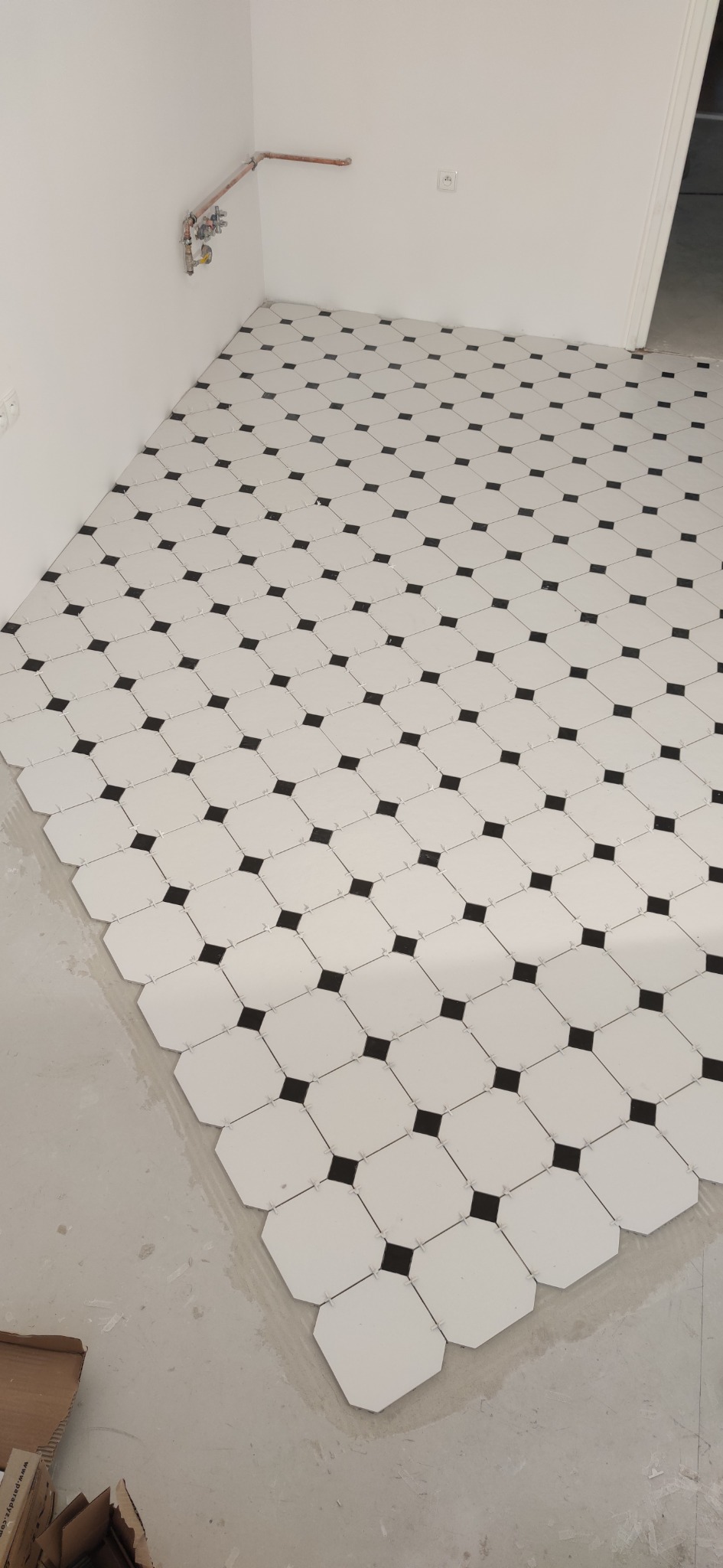 Układanie białych, ośmiokątnych płytek z czarnymi kwadratami w narożach, widoczne krzyżyki dystansowe, rury miedziane na ścianie
