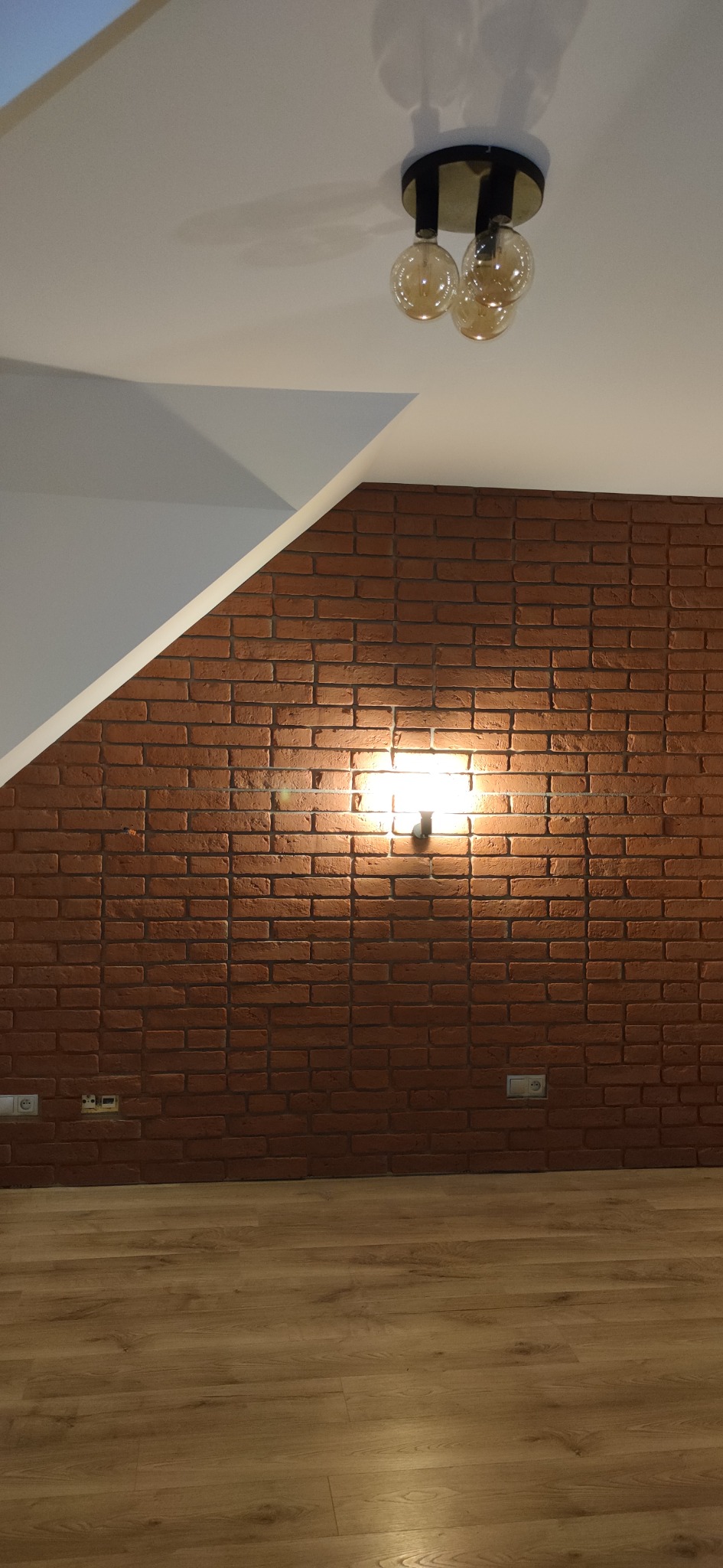 Ceglana ściana z oświetleniem punktowym i drewnianą podłogą, widoczne gniazdka elektryczne, sufit ze skośnym elementem i lampa wisząca z żarówkami Edison.