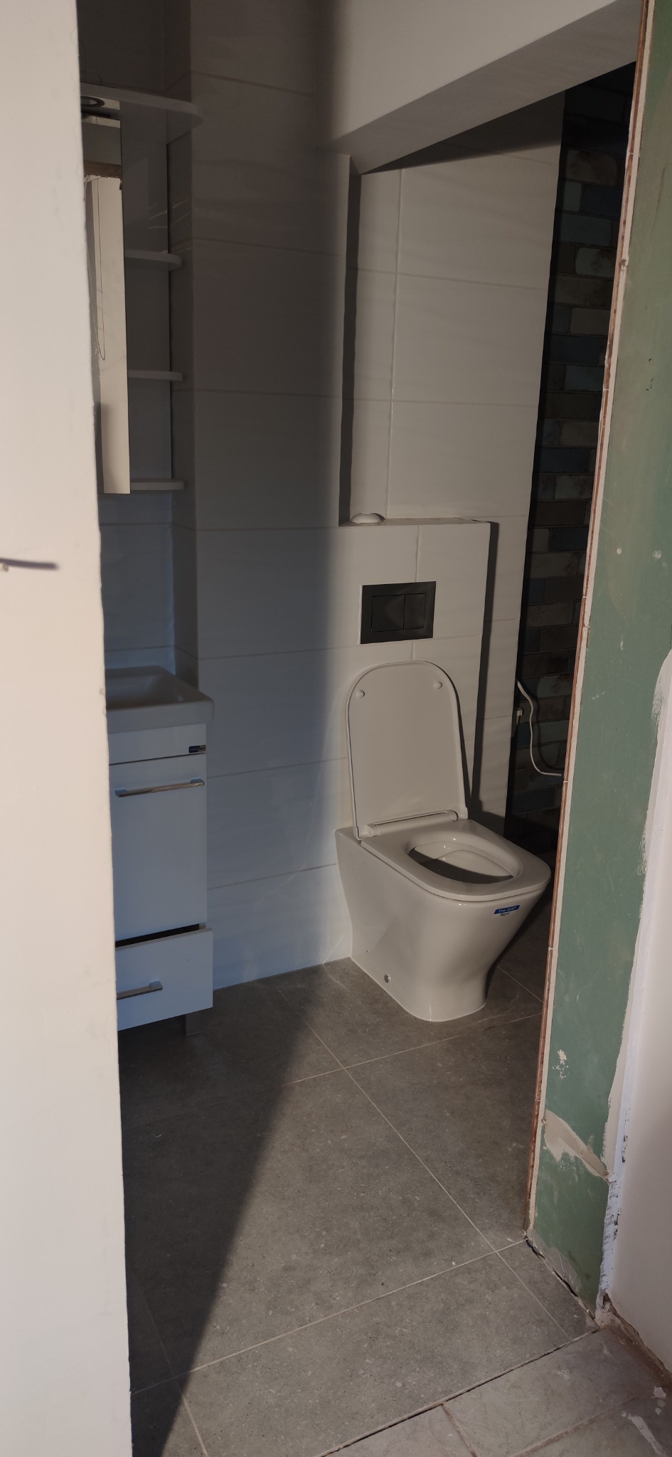 Nowa, biała toaleta typu compact z czarnym przyciskiem spłukującym, zamontowana w łazience z szarymi płytkami podłogowymi i częściowo wykończonymi ścianami, widok zza niedokończonej futryny.