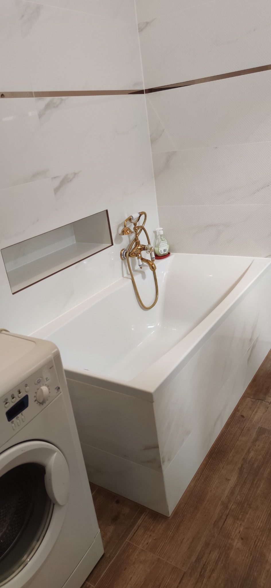 Biała wanna z armaturą w stylu retro, wbudowana w ścianę półka i pralka w łazience wykończonej płytkami imitującymi marmur.