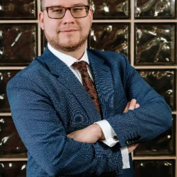 Portret mężczyzny w niebieskim garniturze i okularach, z rękami skrzyżowanymi na tle ściany z luksferów.