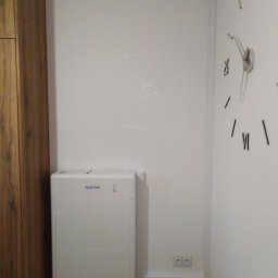 ASFROS SP. Z O.O. - Biały, kompaktowy magazyn energii Sofar, umieszczony w rogu pokoju, obok drewnianej szafy i ściany z minimalistycznym zegarem.