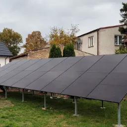 Instalacja paneli słonecznych na metalowej konstrukcji w ogrodzie, z widocznymi domami w tle, pochmurne niebo.