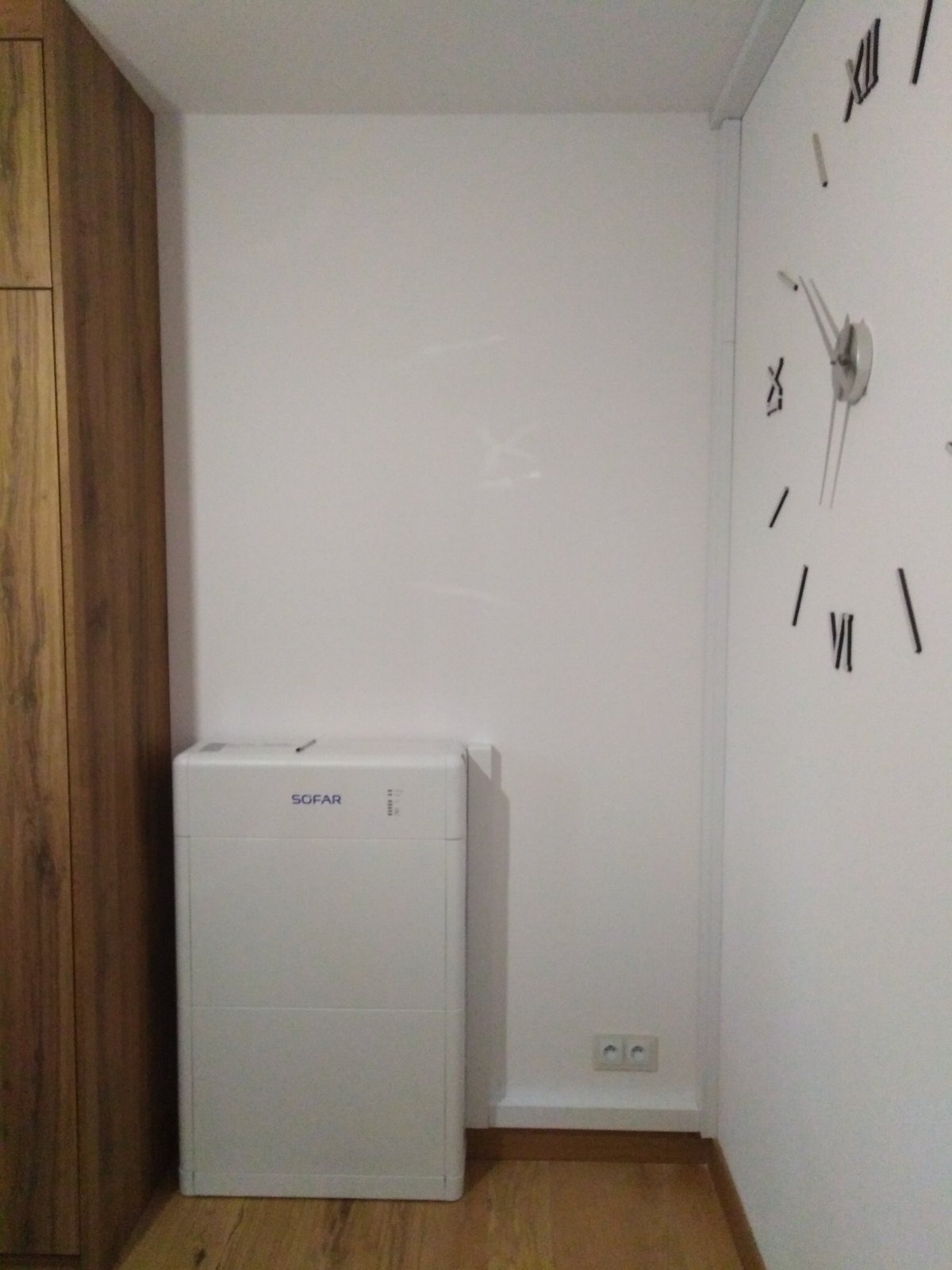 Biały, kompaktowy magazyn energii Sofar, umieszczony w rogu pokoju, obok drewnianej szafy i ściany z minimalistycznym zegarem.
