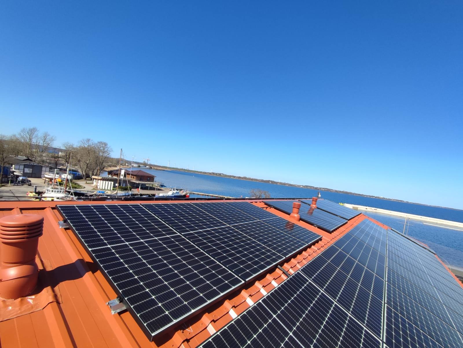 Panele fotowoltaiczne na pomarańczowym dachu z widokiem na morze i zabudowania portowe w tle, błękitne niebo.