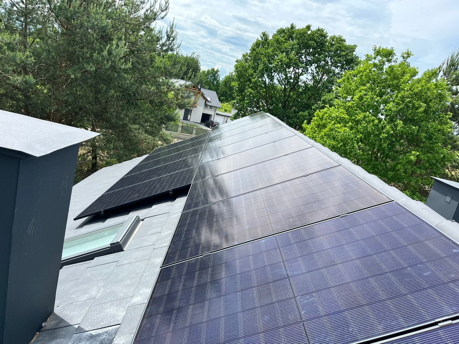 Panele słoneczne na dachu domu jednorodzinnego z szarymi dachówkami, widok z boku, z kominem i oknem dachowym, drzewa w tle.