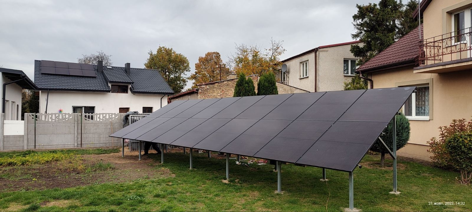 Naziemna instalacja paneli słonecznych na metalowej konstrukcji w ogrodzie, widoczne domy w tle, pochmurne niebo.