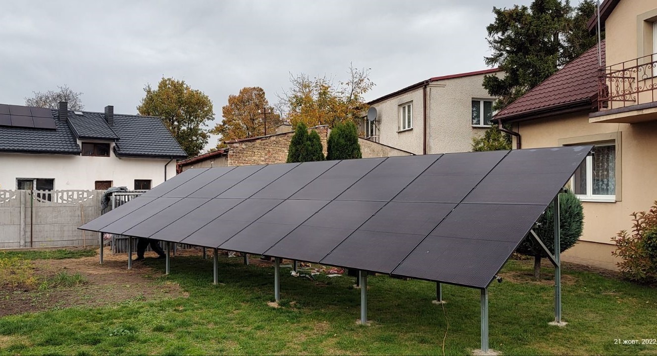 Instalacja paneli słonecznych na metalowej konstrukcji w ogrodzie, z widocznymi domami w tle, pochmurne niebo.