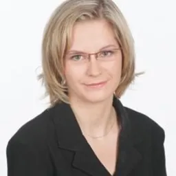 Portret kobiety w okularach, z blond włosami do ramion, ubrana w czarną marynarkę, na jasnym tle.