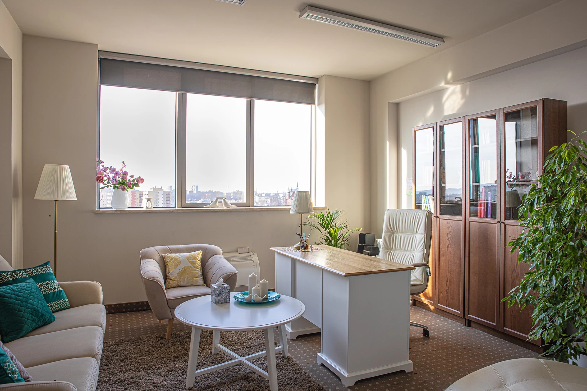 Jasne, przestronne wnętrze gabinetu z dużym oknem, biurkiem z drewnianym blatem, wygodnym fotelem i sofą z dekoracyjnymi poduszkami, całość utrzymana w ciepłej, stonowanej kolorystyce.