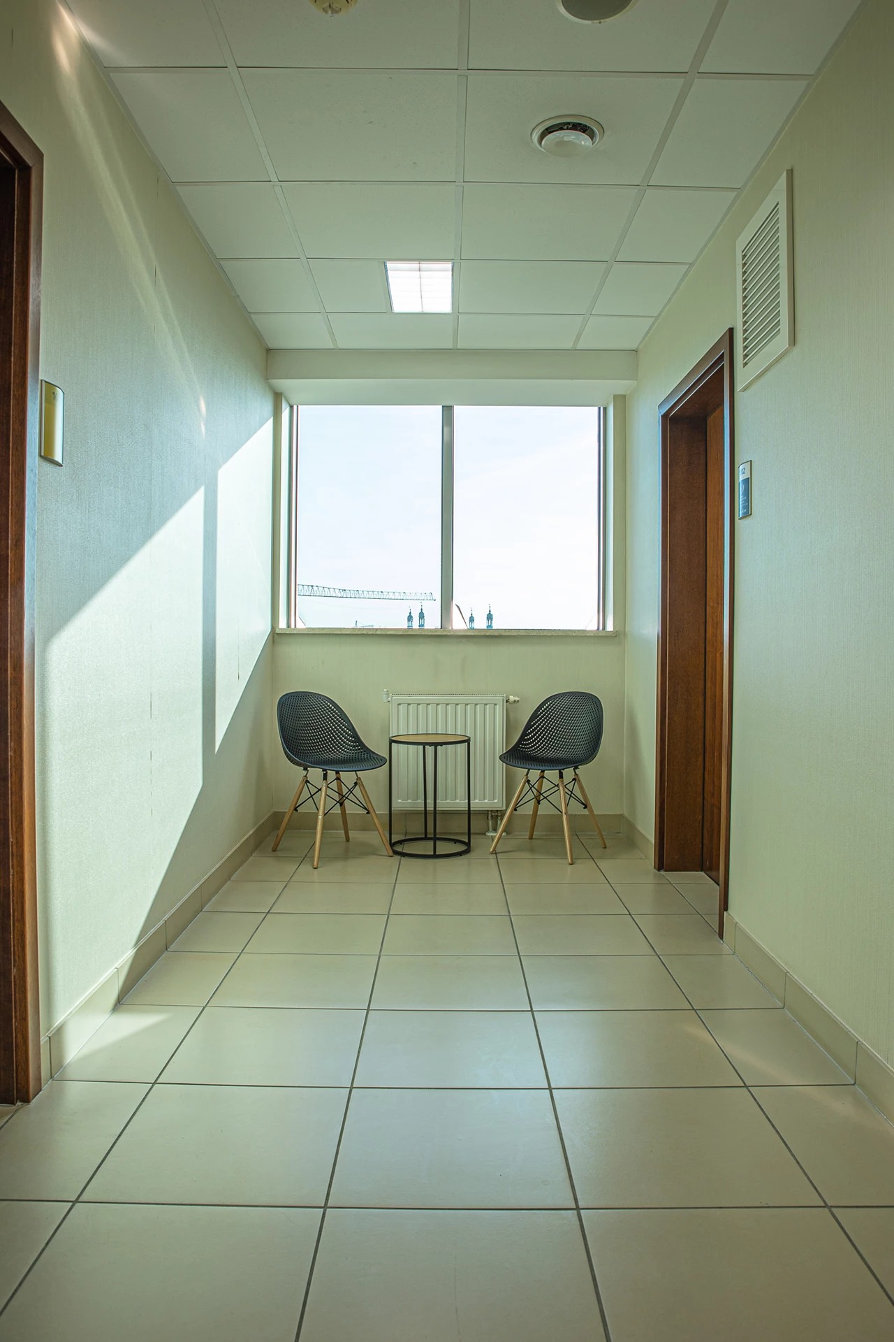 Jasny korytarz z dwoma krzesłami i stolikiem przy oknie, widok na budowę za oknem.