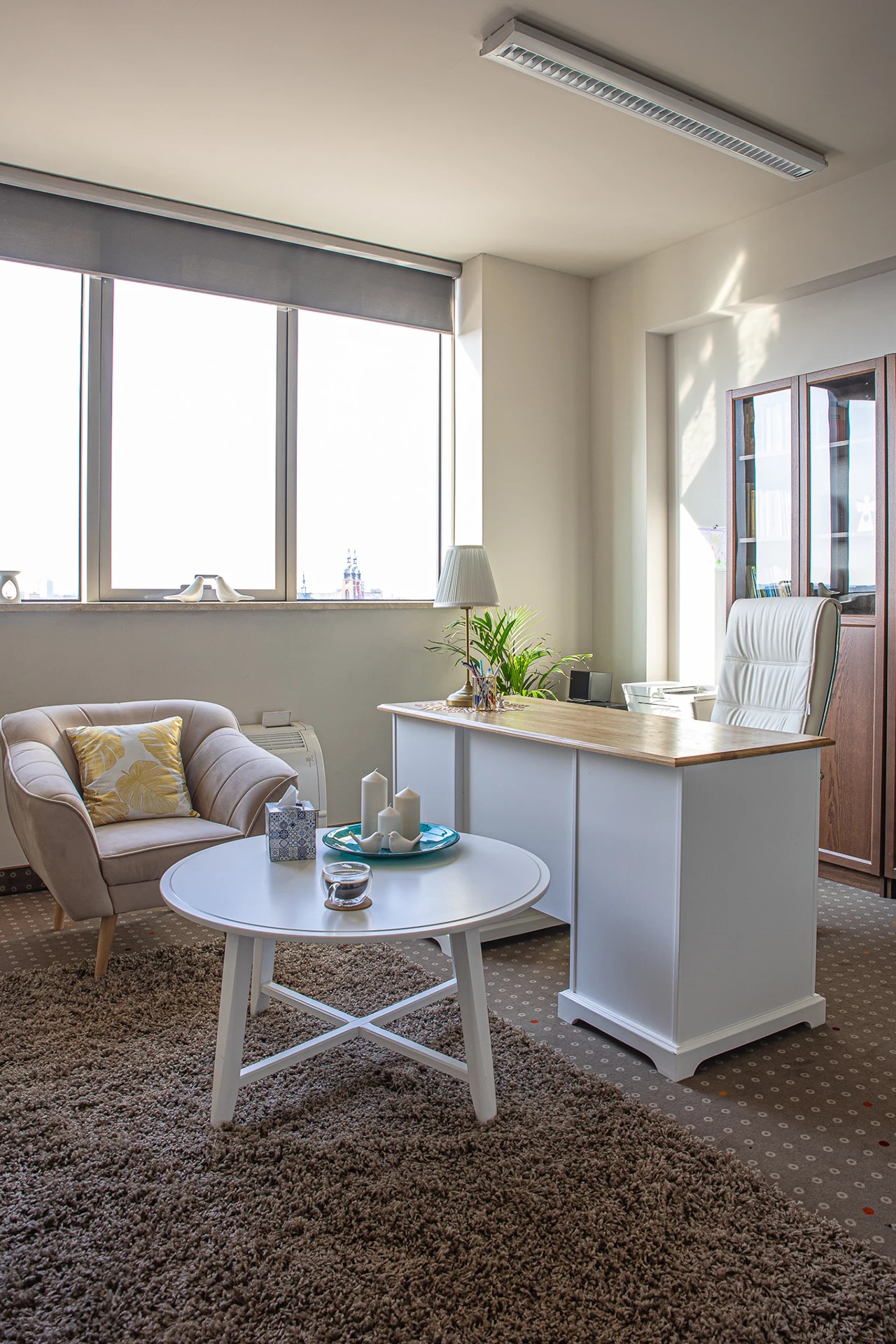 Jasne, przestronne biuro z dużym oknem, beżowym fotelem z poduszką, białym stolikiem kawowym z dekoracjami, biurkiem z drewnianym blatem i białymi szafkami oraz skórzanym krzesłem biurowym.