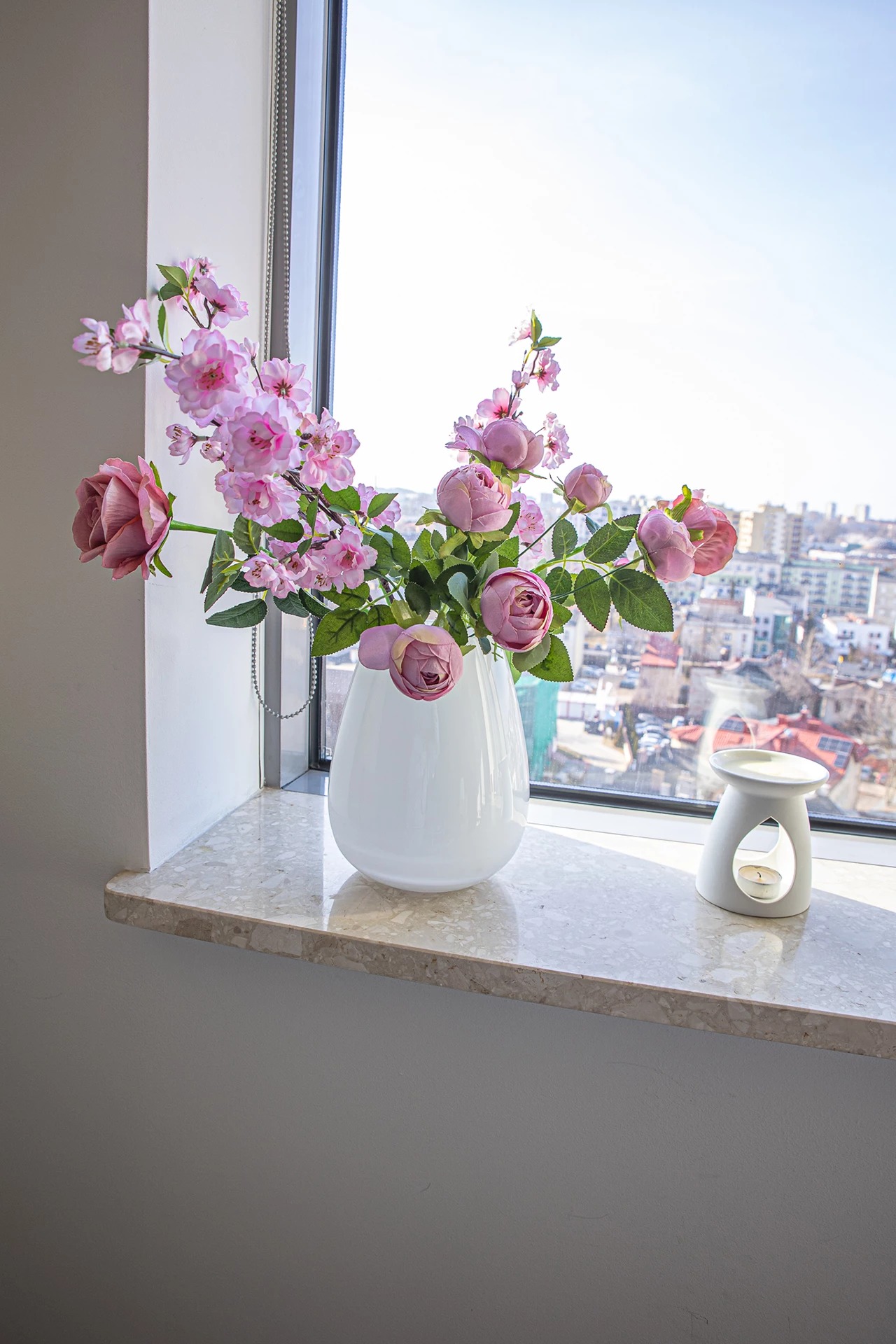 Bukiet różowych kwiatów w białym wazonie na marmurowym parapecie okiennym z widokiem na miasto w tle, obok ceramiczny podgrzewacz do olejków eterycznych.